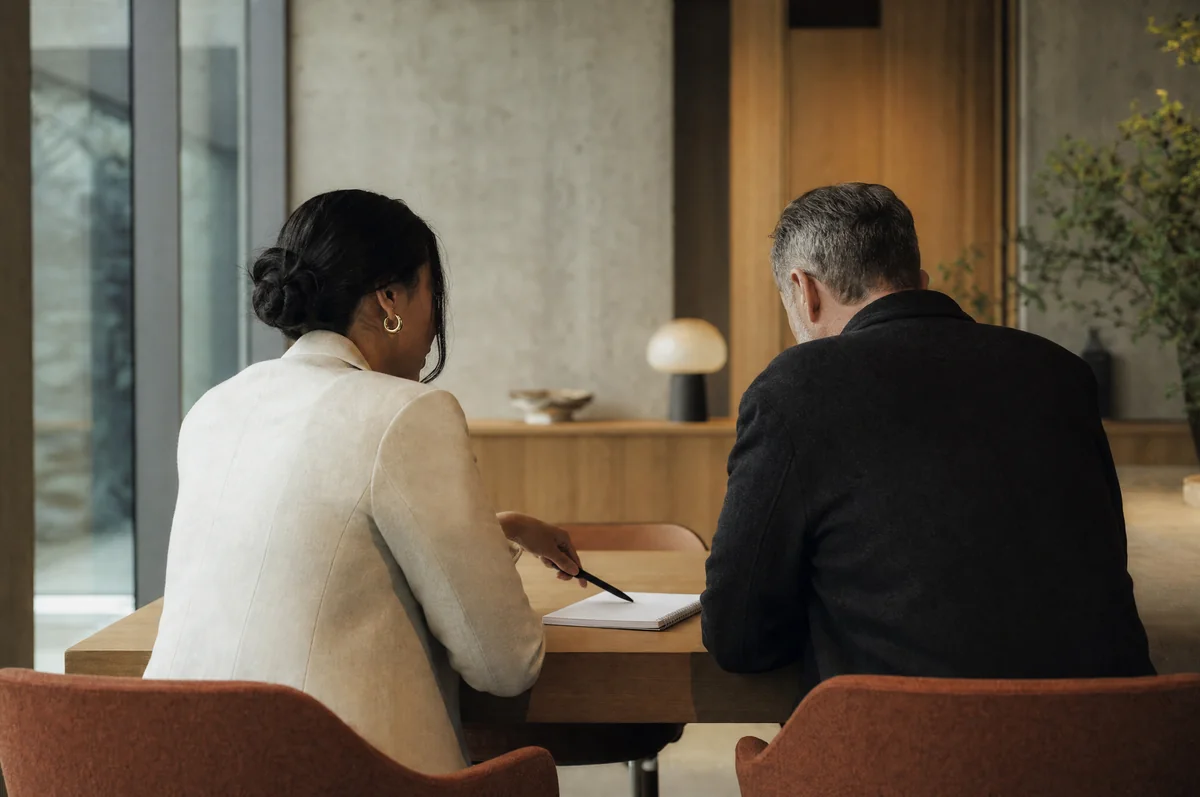 An adviser and a client, seen from behind, sitting side by side at a timber table in a modern Sydney office. The adviser points to a notepad.