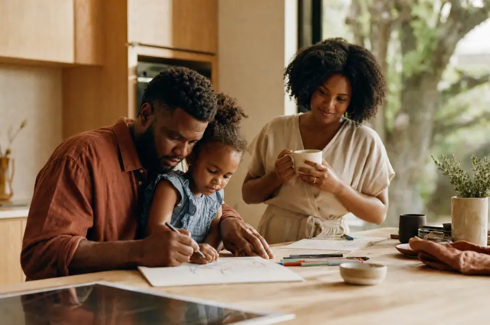 A family at a kitchen table, a parent helping a young child with drawing while another adult looks on.